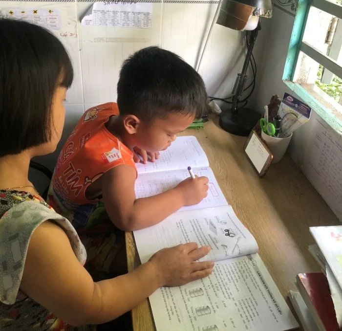 Bé trai học tập viết bài trong vở có mẹ hướng dẫn tại bàn học